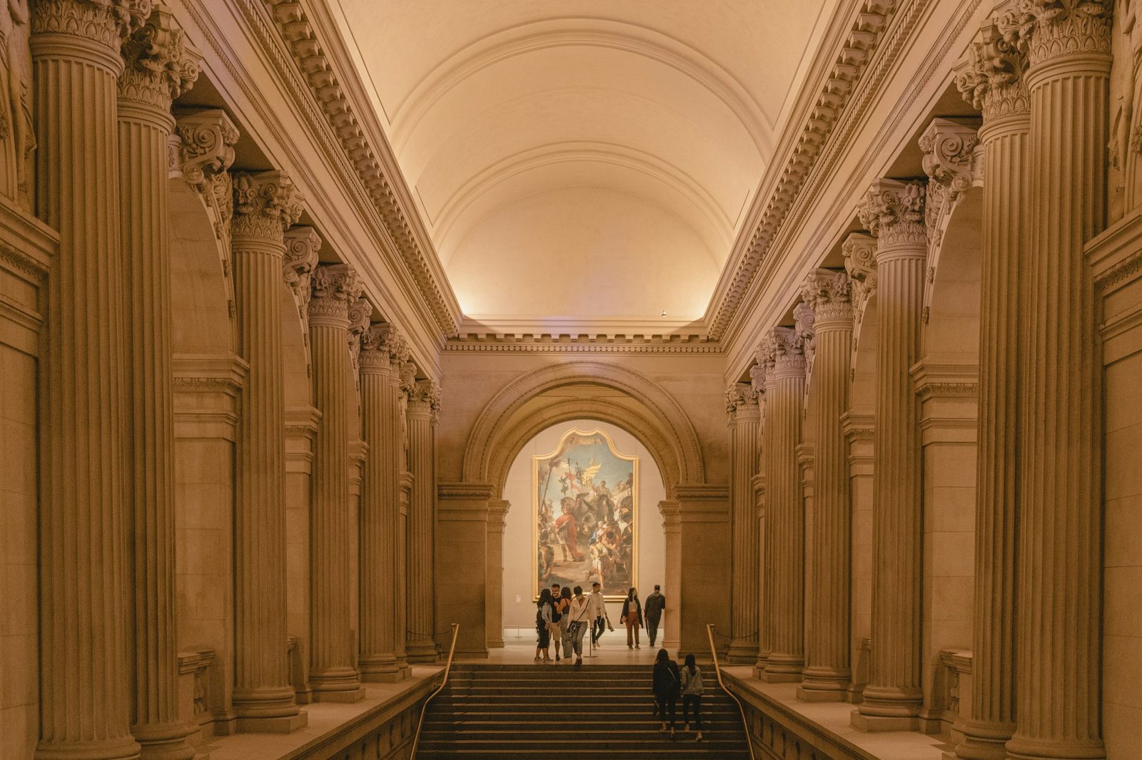 A columned stone hall with a staircase and a large painting at the far end