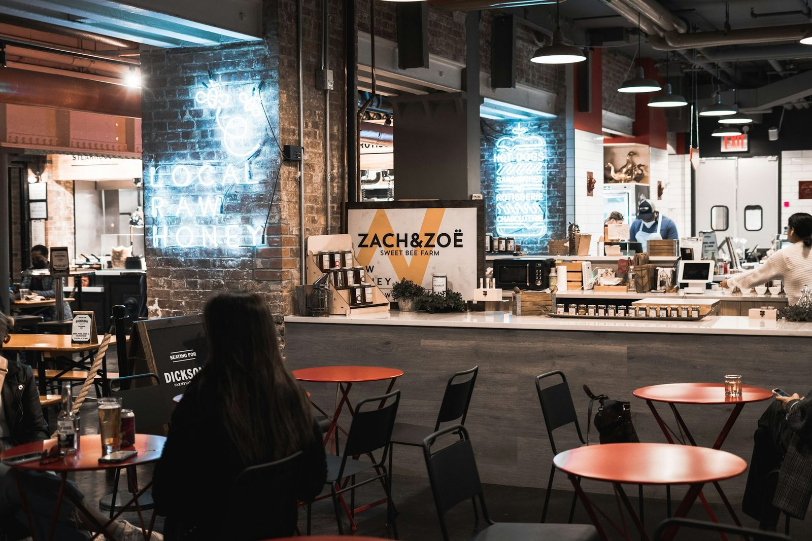 A busy cafe with red tables, an open kitchen counter and neon signs on a brick wall