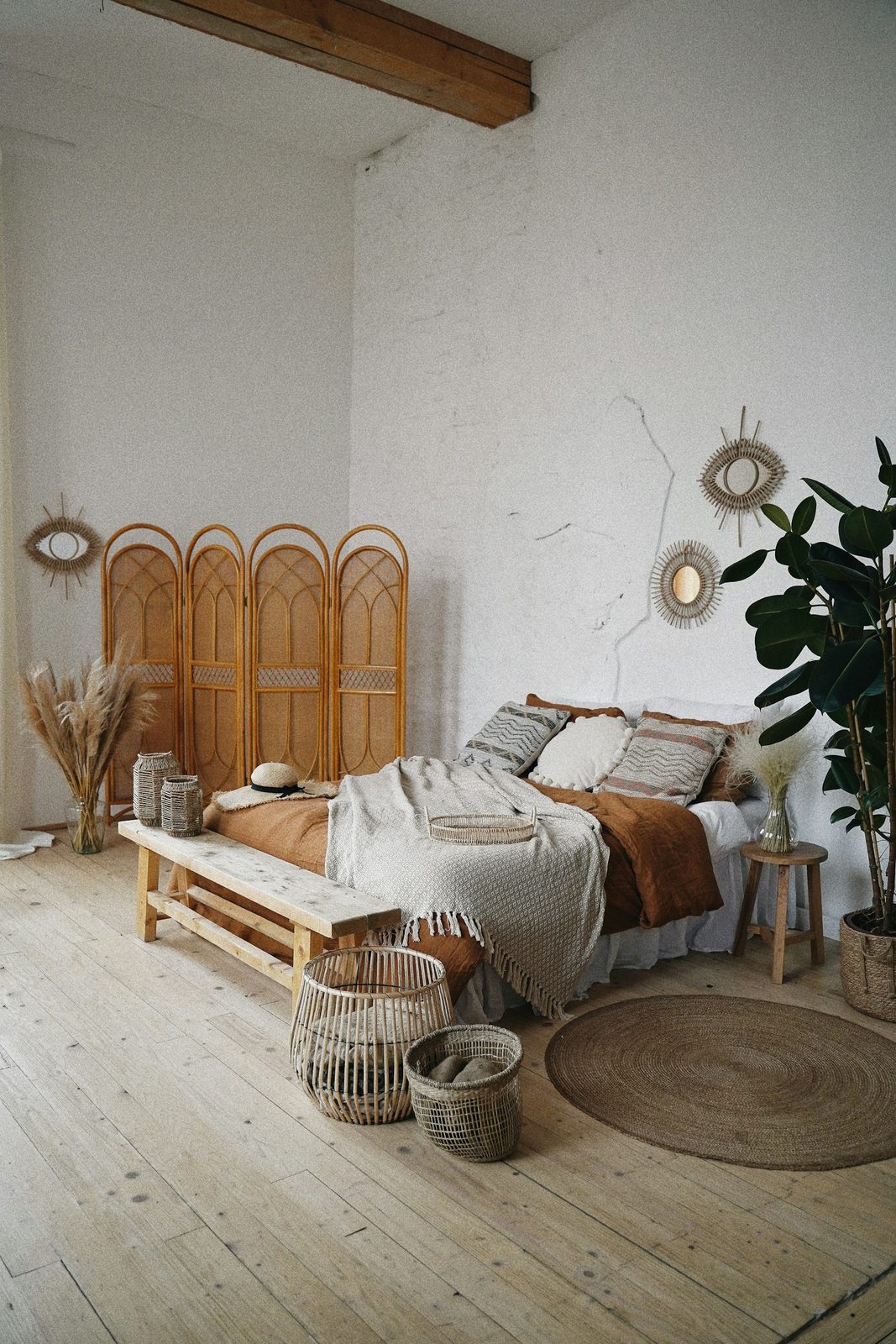 A bed with a woven headboard screen, a wooden bench, a jute rug and a large plant in a white room with a dark ceiling beam