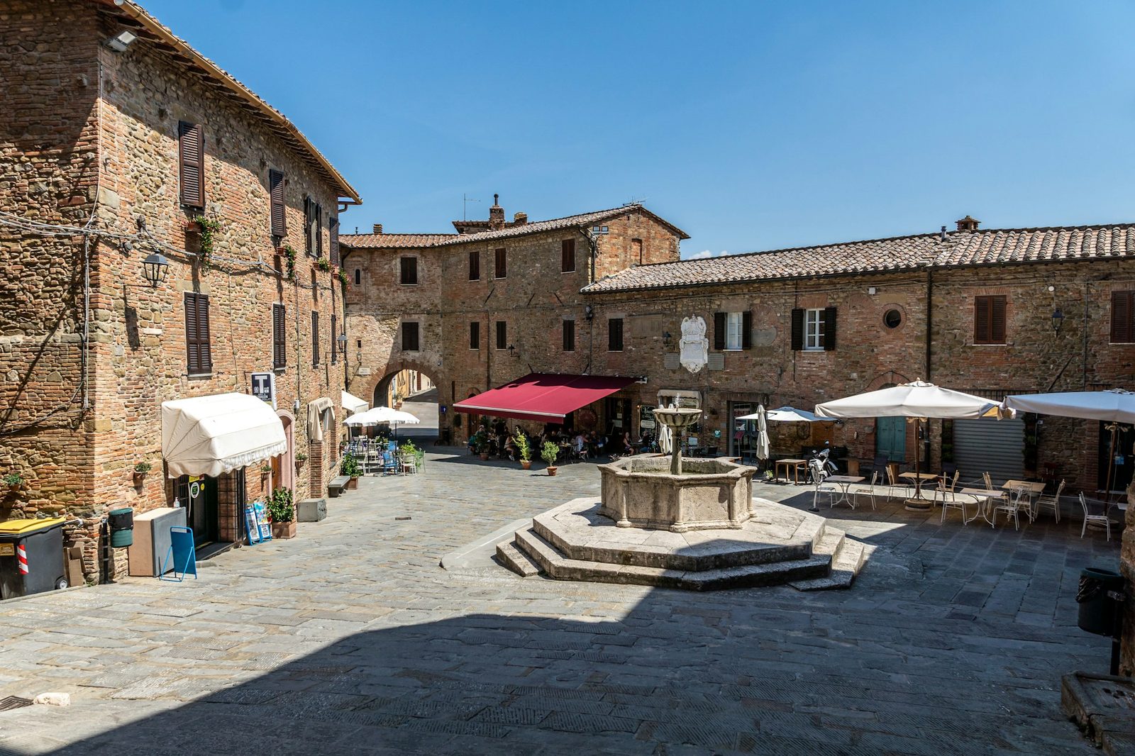 A stone square with a well-head fountain, awnings and café tables in the sun