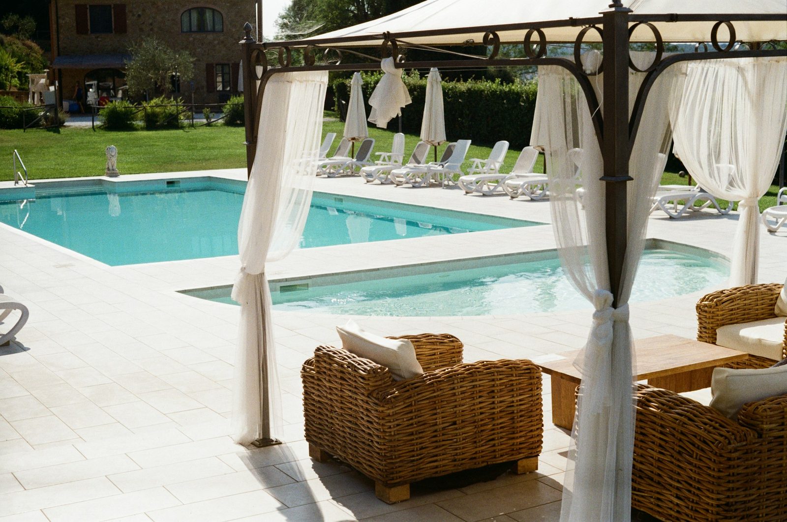 A pergola with white drapes and wicker armchairs beside a pool, with loungers on the lawn beyond