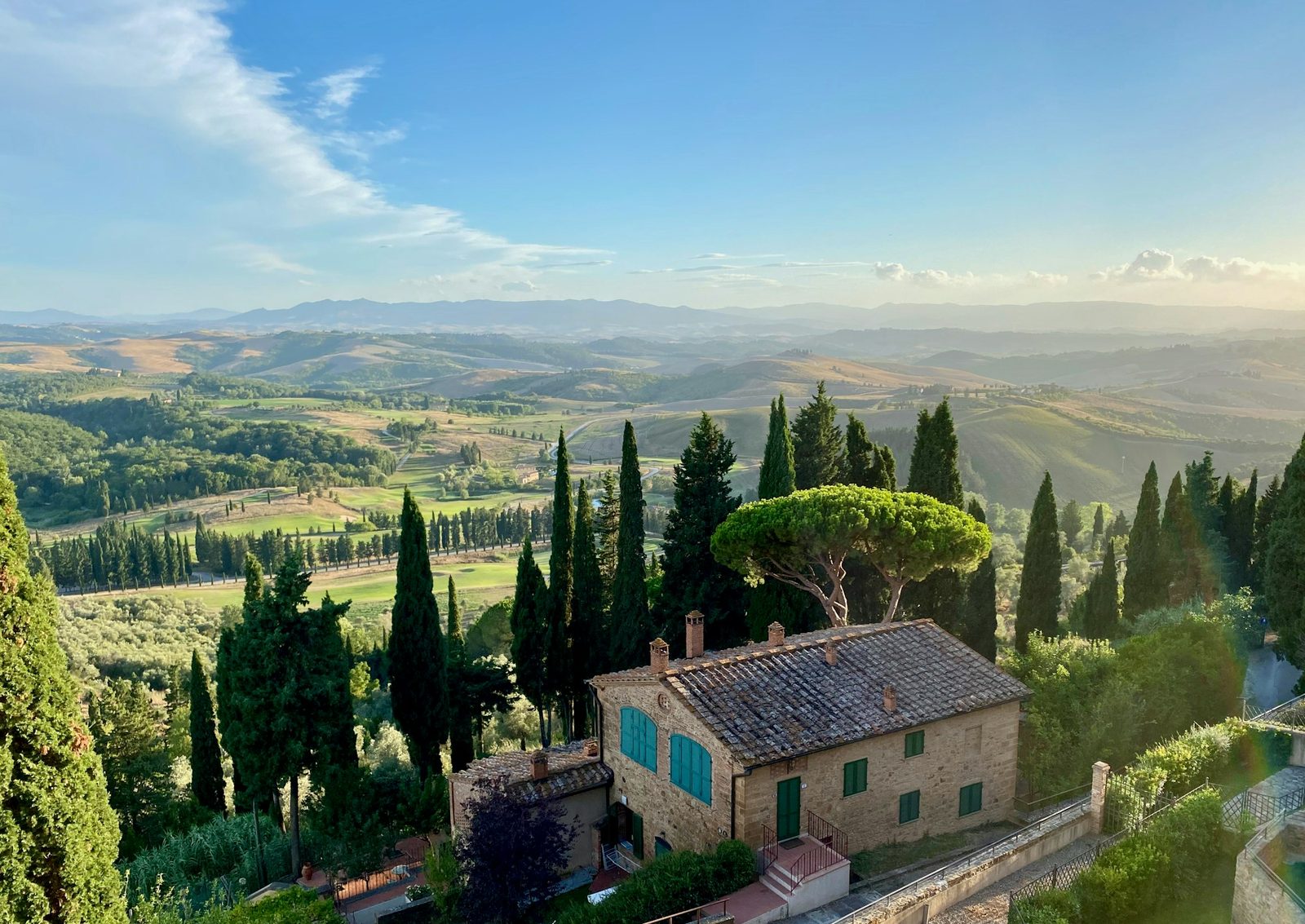 Looking down on a stone farmhouse with a tiled roof and blue-green shutters, cypresses and an umbrella pine in front, hills fading into the distance