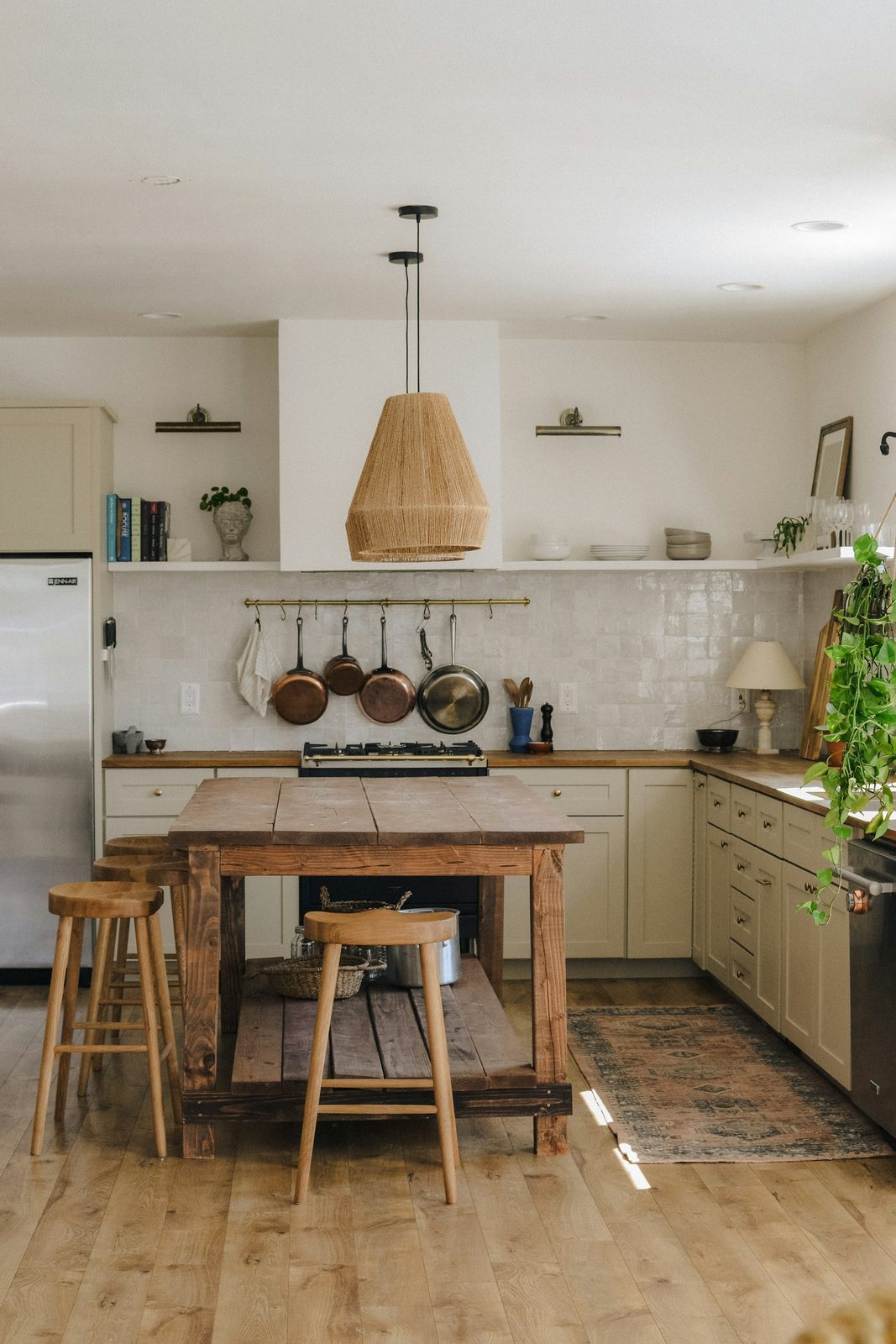 A kitchen with white tiled walls, a long timber table with stools, a woven pendant lamp and copper pans on a rail