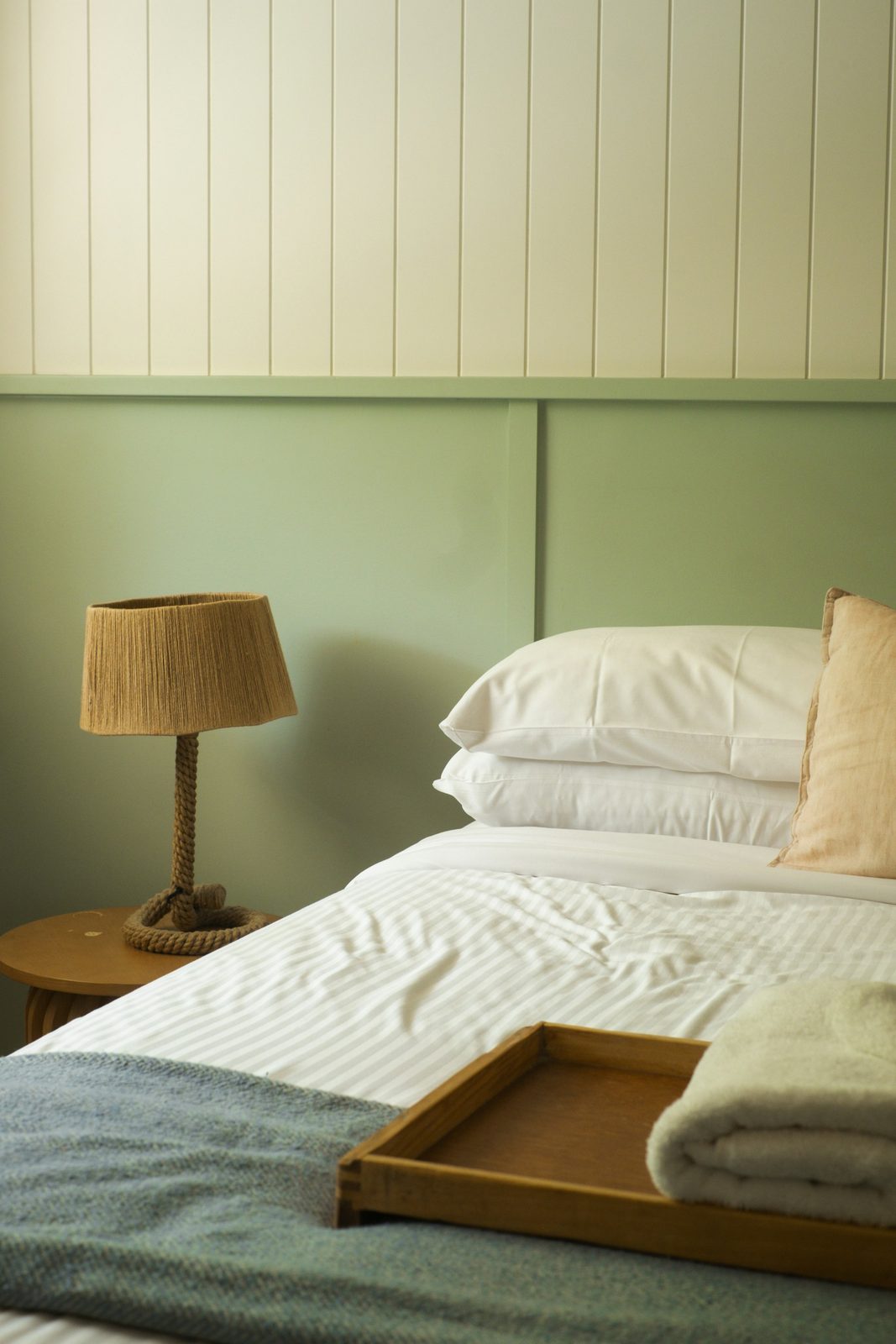 A bed with white linen, a woven lamp and a wooden tray, against a sage green and white panelled wall.