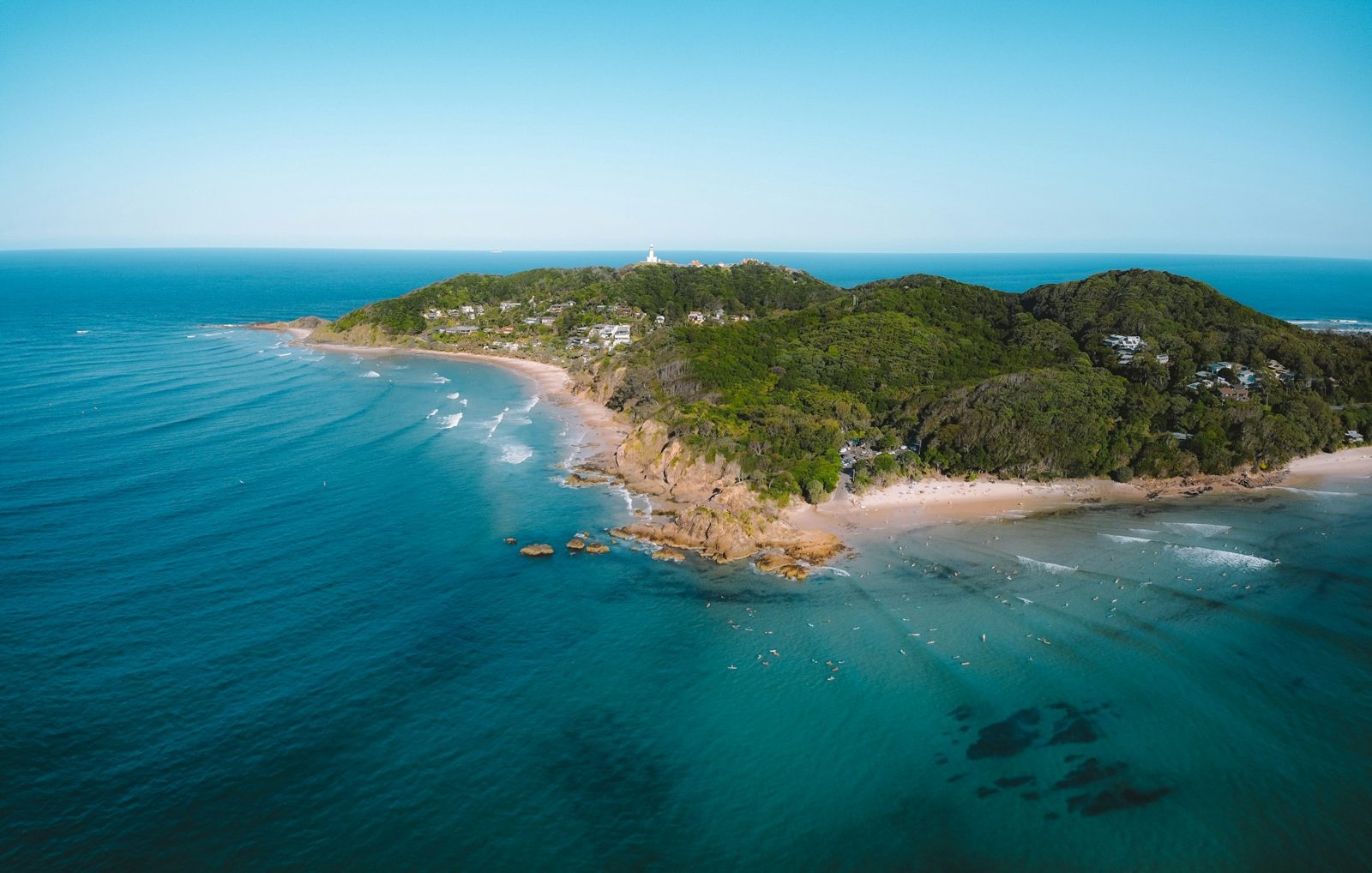 An aerial view of a green headland with a small lighthouse on top, a rocky shore and turquoise water.