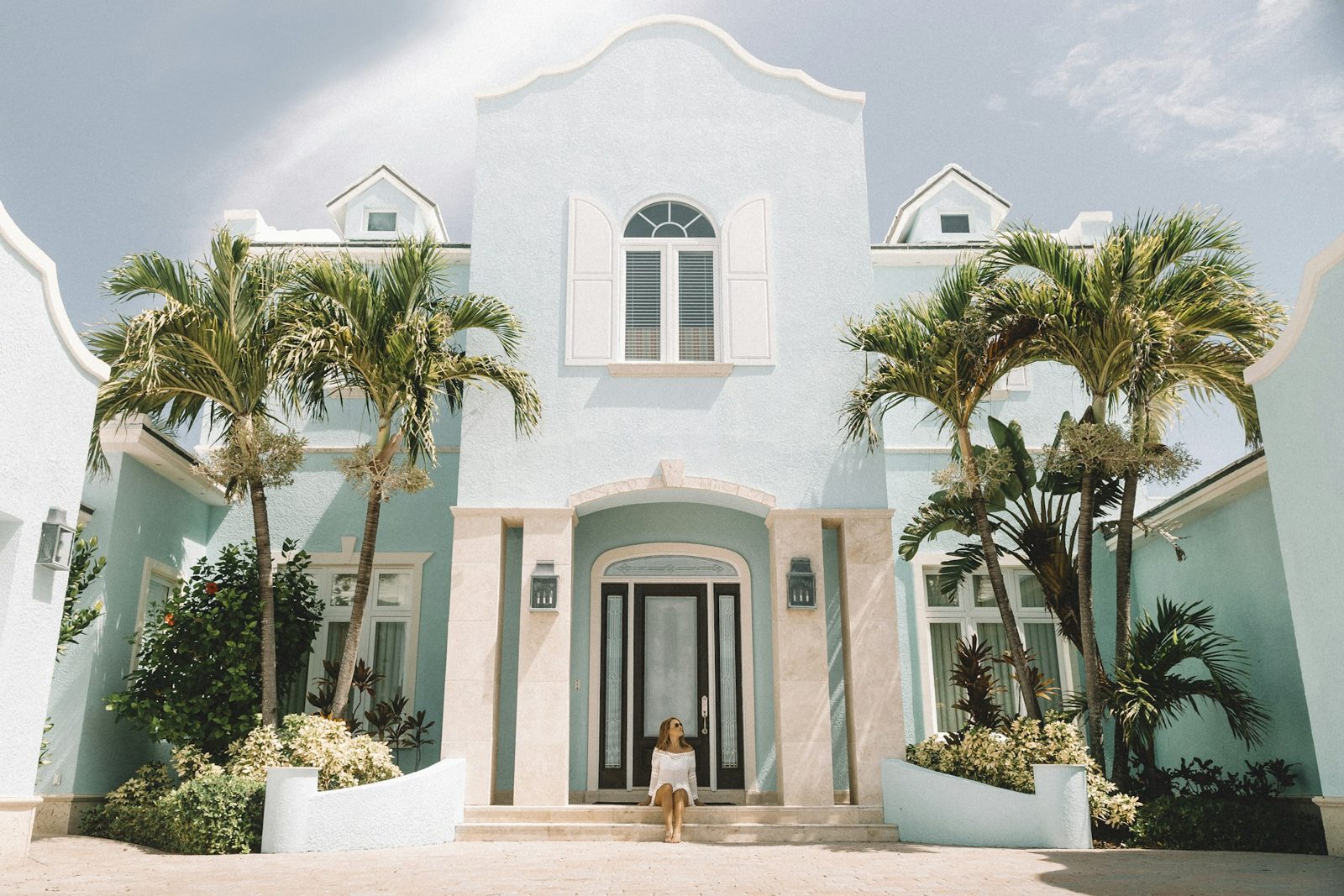 The front of a pale blue and white house with arched windows, a shuttered gable and a palm tree on each side of the front door.