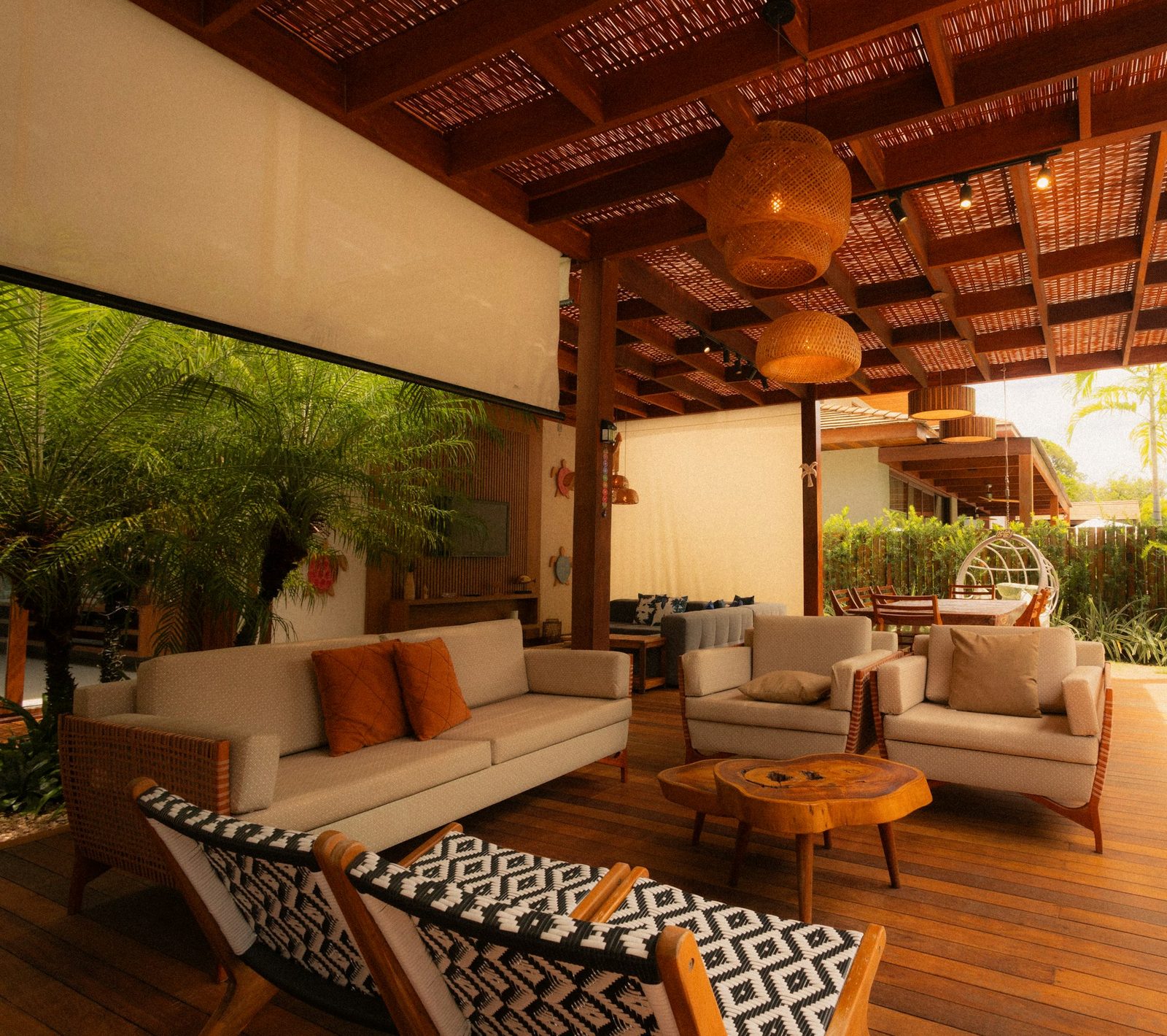 A lounge under a high timber ceiling, with cane-framed sofas, patterned armchairs and tropical plants at the open side.