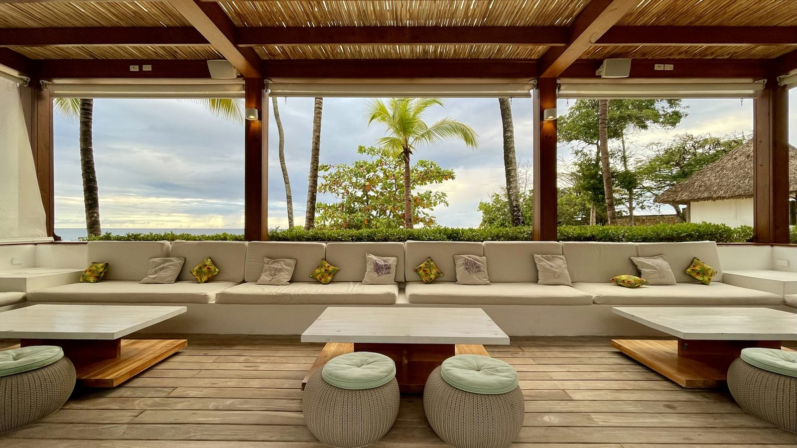 A covered terrace with a long cushioned sofa, low stone tables and round woven stools, open to palms.