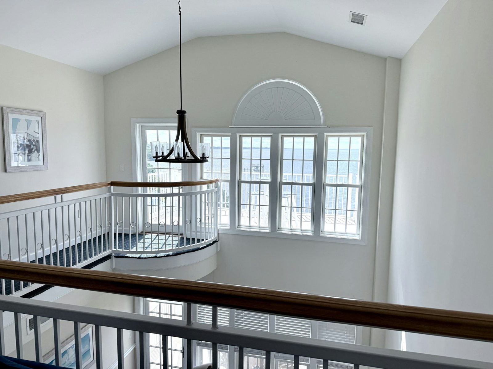 A staircase with white balustrades curving beneath a tall arched window and a black chandelier.