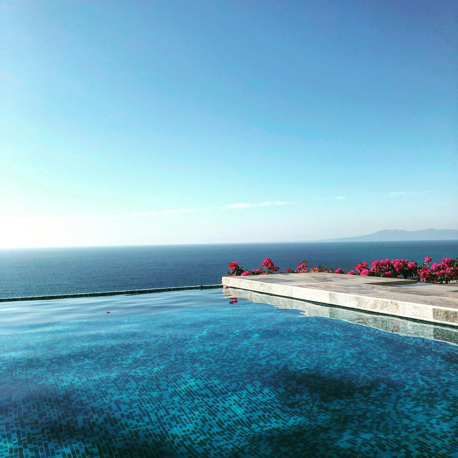 An infinity-edge pool with a tiled floor, pink bougainvillea along the far edge and the sea beyond.