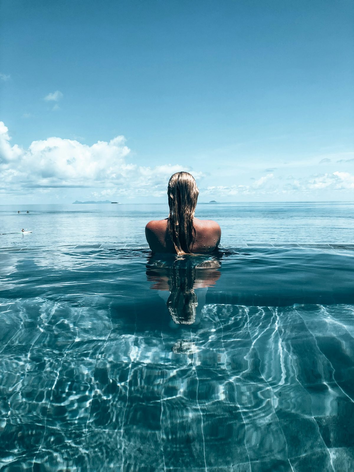 A person standing in clear water at the edge of an infinity pool, looking out to sea.