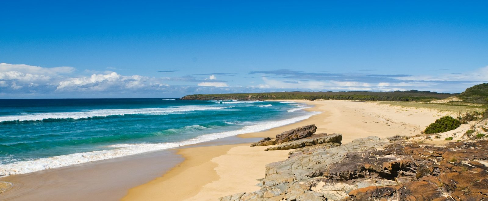 A wide sandy beach curving away to a headland, with rocks in the foreground and a deep blue sea.