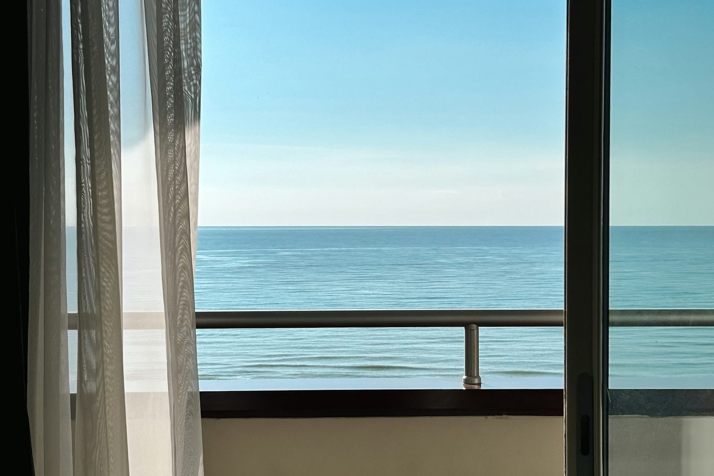A sheer curtain and an open glass door onto a balcony, with the sea and a clear blue sky beyond the railing