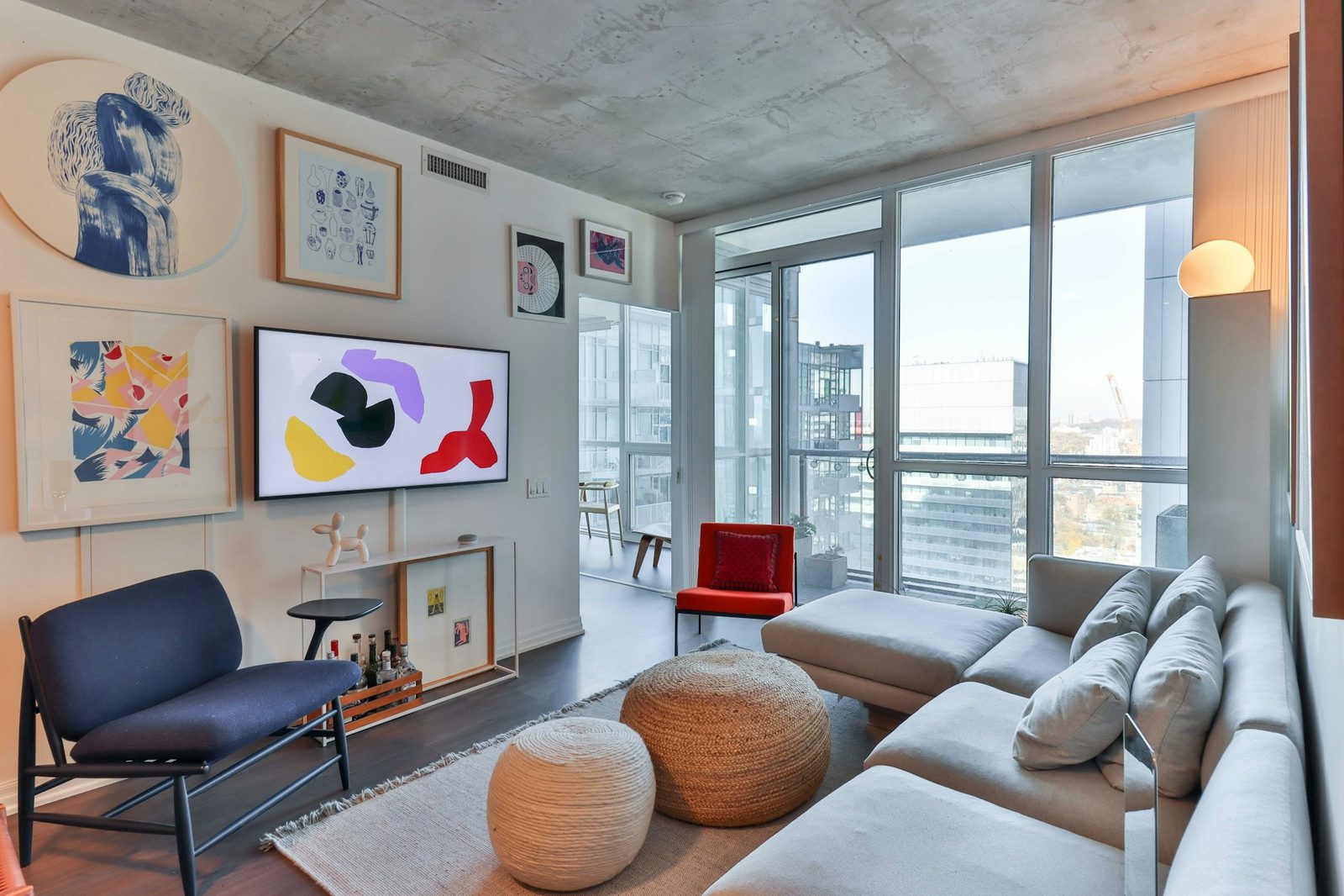 A living room with a grey sectional sofa, a blue armchair, framed prints on the wall and tall windows onto a city view