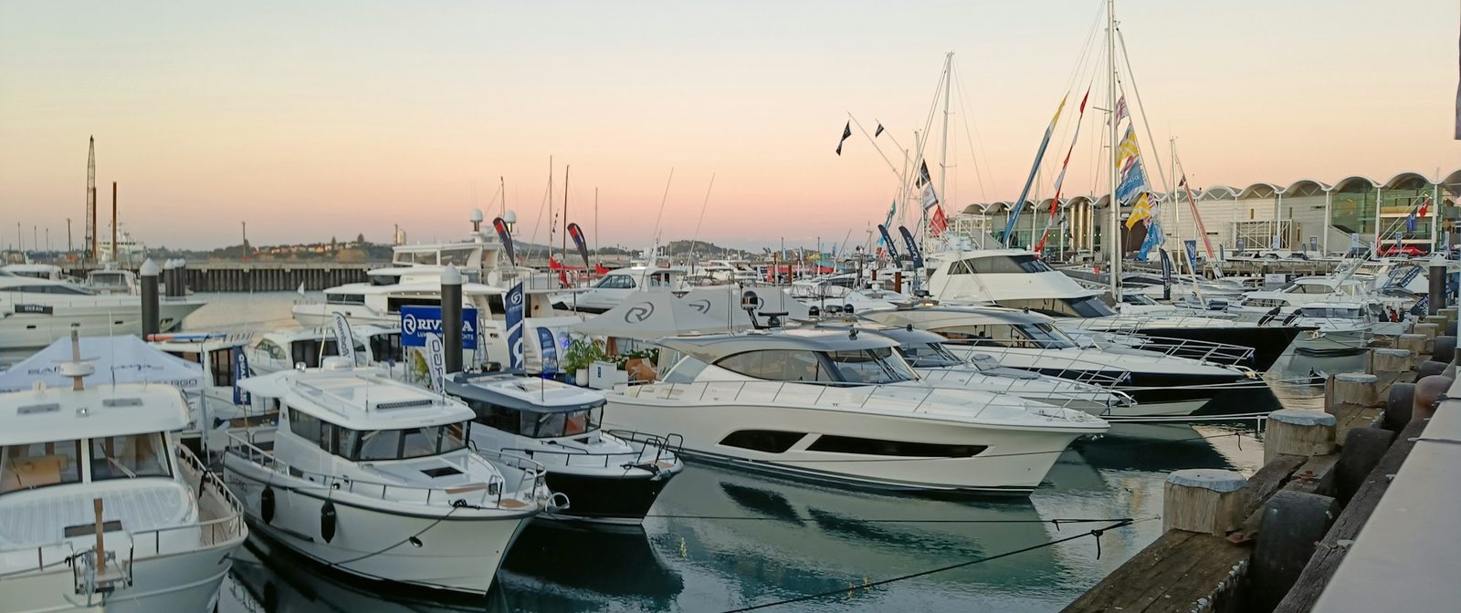 Motor yachts and cruisers moored in a marina at sunset, with flags and masts along the far quay